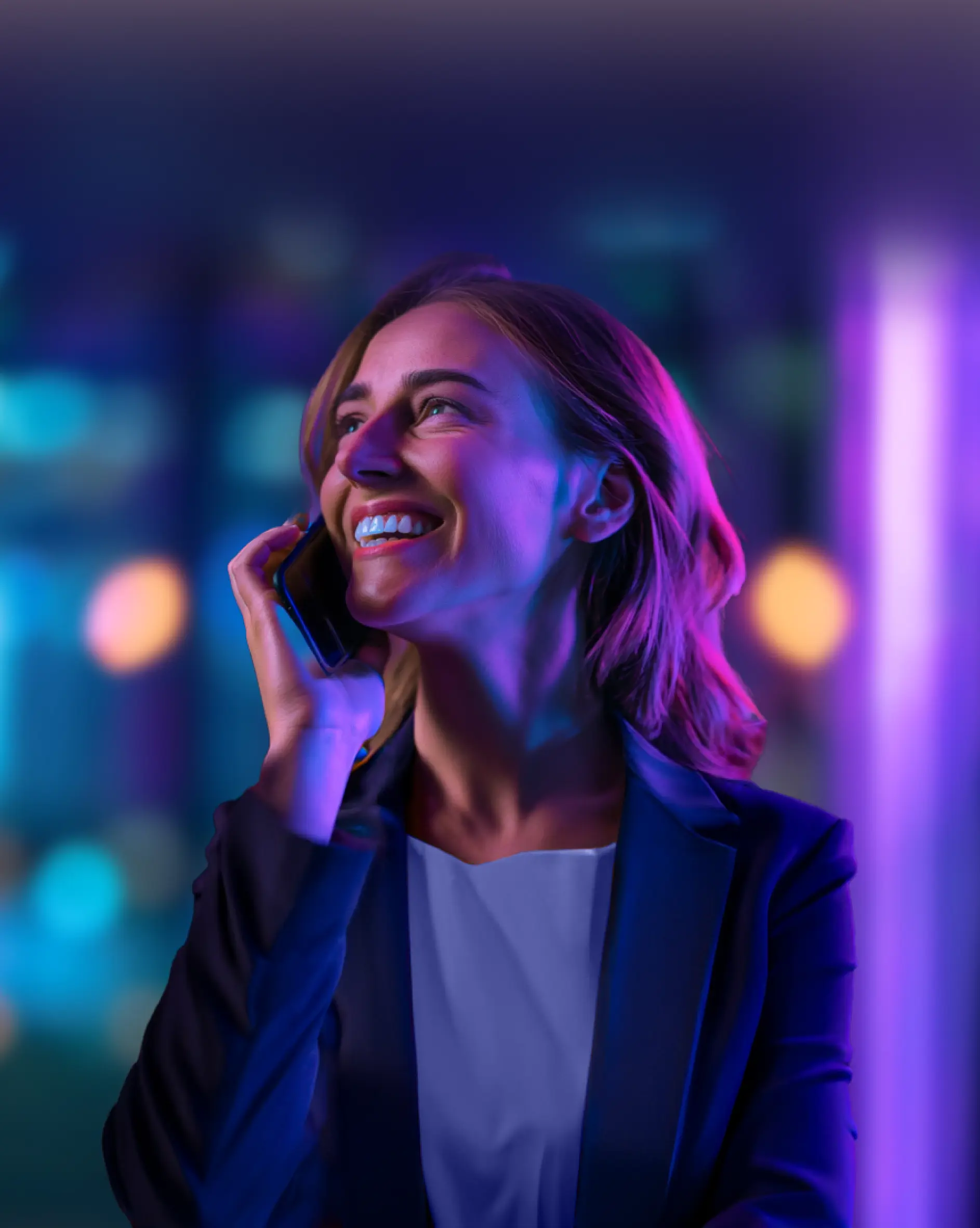 Woman in a business suit smiling and talking on a smartphone with a blurred background.