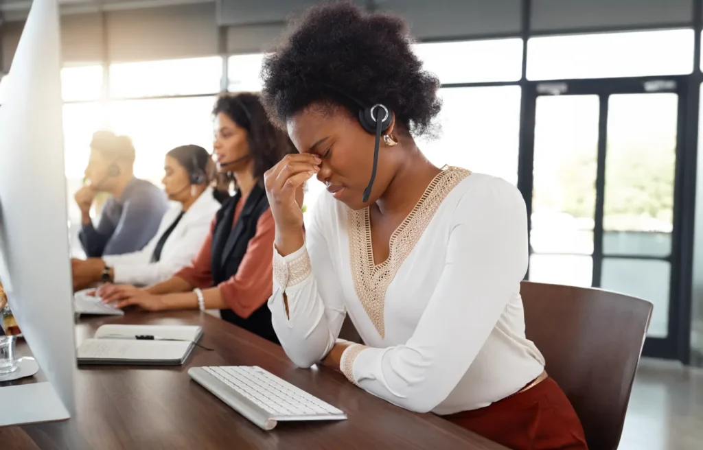 Woman wearing a headset sitting at a desk, looking stressed with her hand on her face, while other employees work at computers in the background.