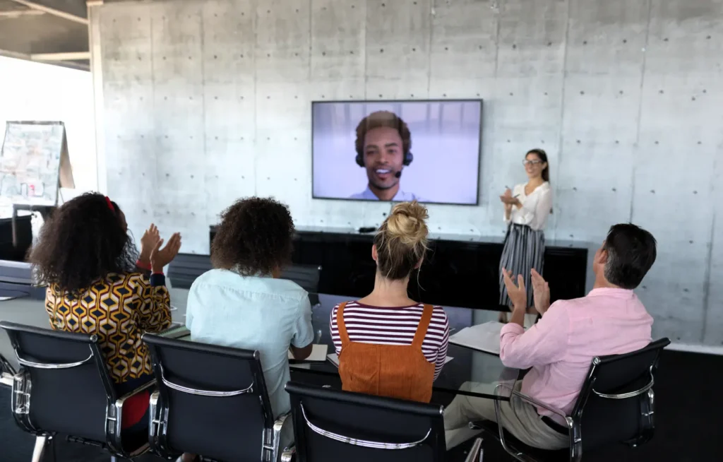 Group of people in a conference room clapping while watching a video call presentation on a large screen.