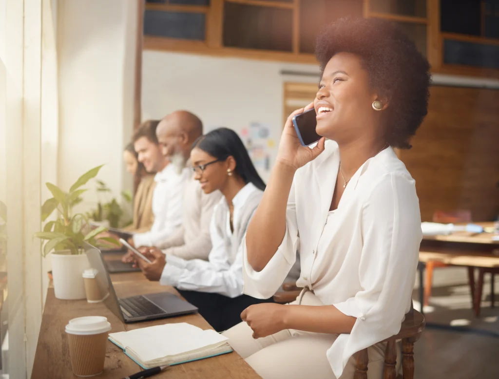 Woman smiling and talking on a smartphone, seated in a shared workspace with colleagues working in the background.