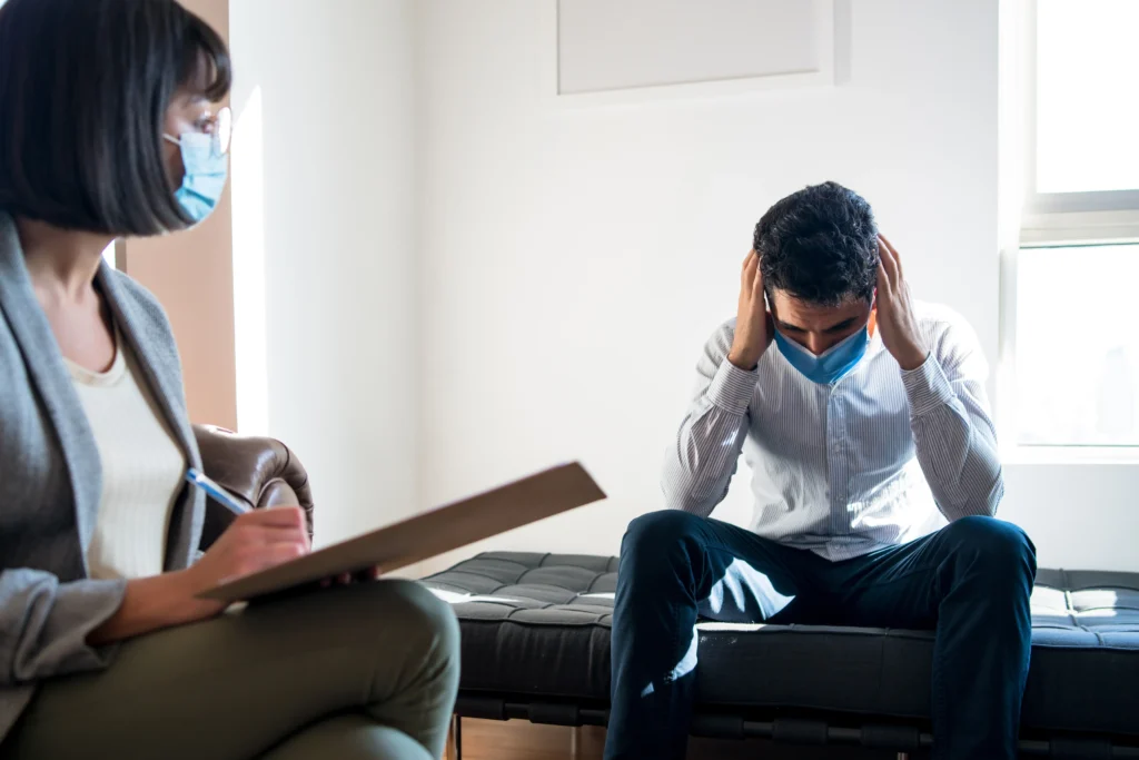 Man wearing a face mask sitting with his head in his hands during a therapy session, while the therapist takes notes.