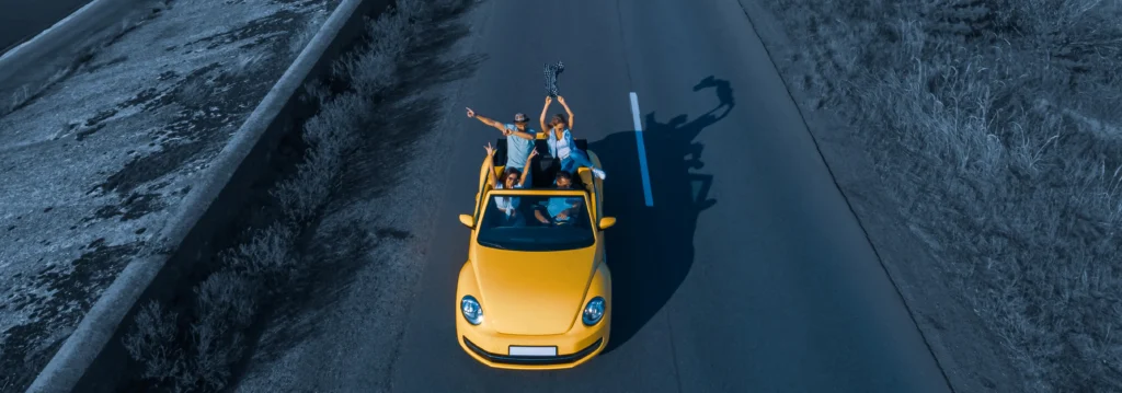 Group of friends enjoying a road trip in a yellow convertible, driving down an open road with arms raised.