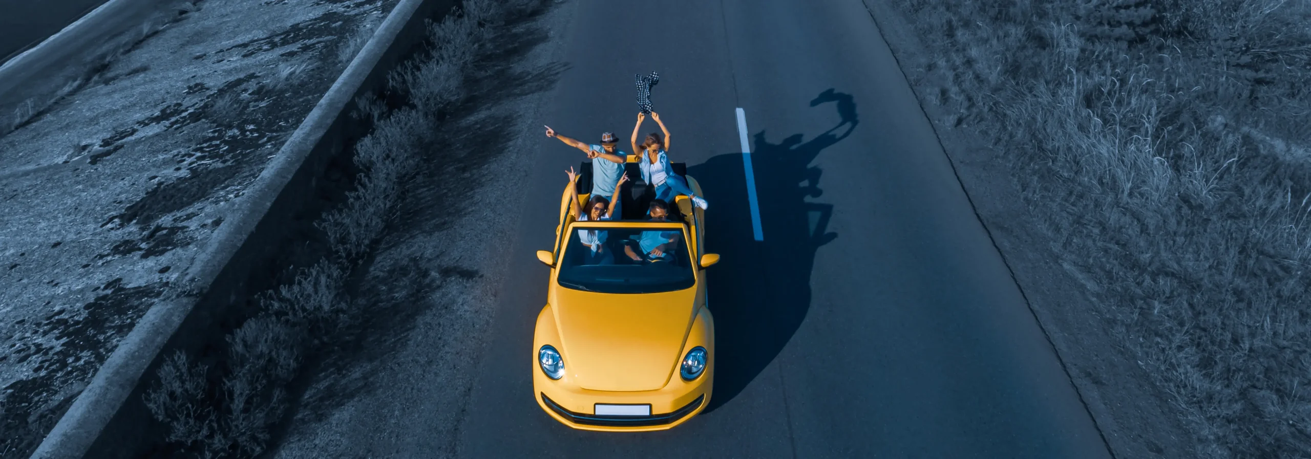 Group of friends enjoying a road trip in a yellow convertible, driving down an open road with arms raised.